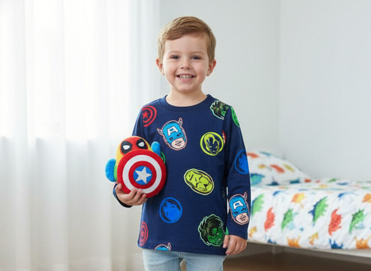 Child wearing a blue shirt with superhero logos, holding a shield-shaped toy, in a bedroom setting.